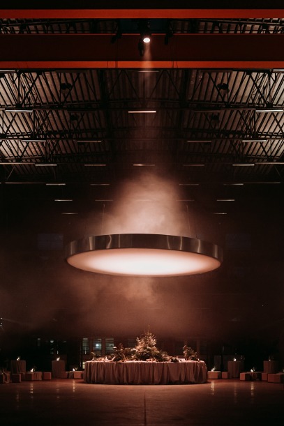 A round table with floral decor sits under a large glowing light in a moody, industrial-style venue